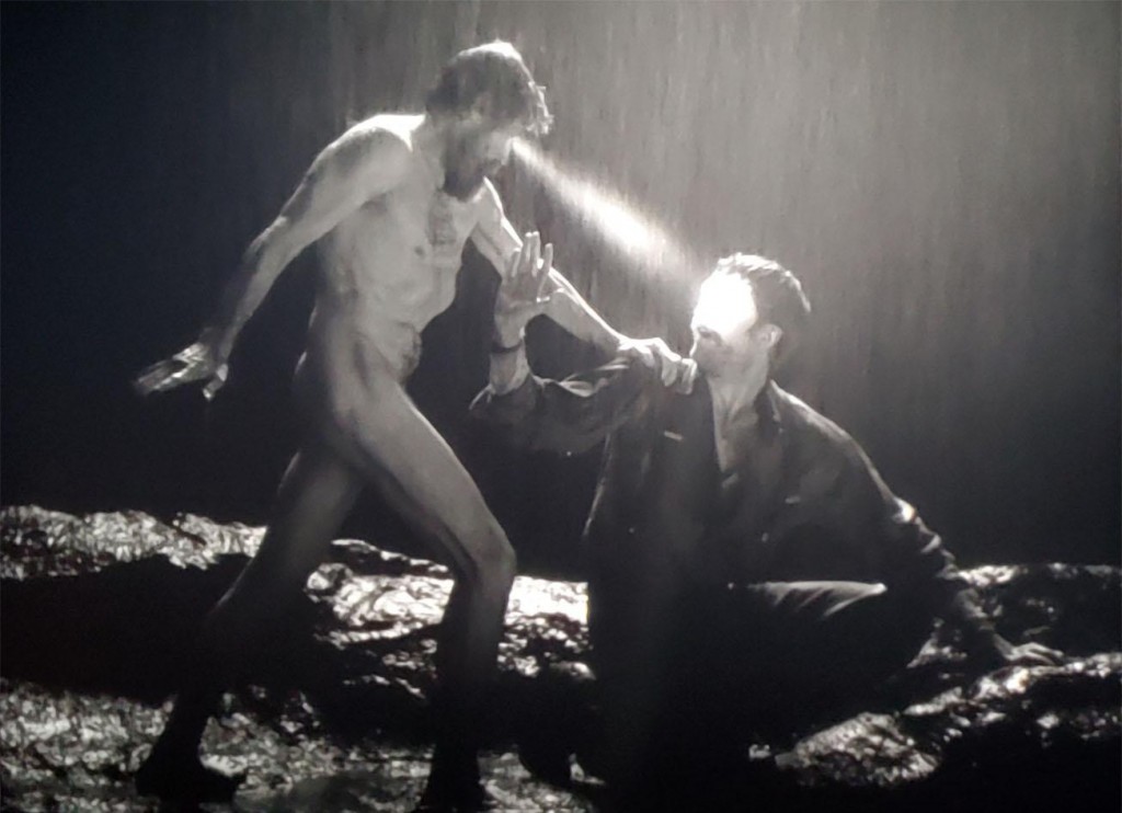 A nude Thomas Wake (Willem Dafoe) uses his lightbeam eyes to hypnotize a kneeling Ephraim Winslow (Robert Pattinson) in the rain.